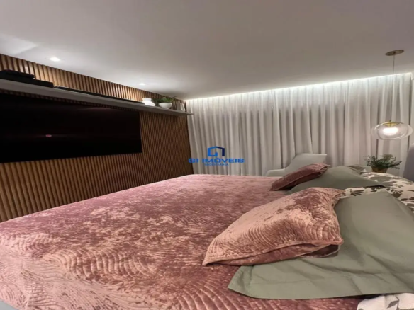 Esta imagem retrata um quarto de dormir com um design contemporâneo e acolhedor, visto a partir de uma perspectiva próxima à base da cama, olhando em direção à cabeceira e à parede lateral. Cama e Roupa de Cama: A cama é o elemento central, coberta por um edredom ou colcha volumosa de veludo em tom rosa queimado ou rosa antigo. A textura do veludo é rica e apresenta um padrão sutil em relevo, possivelmente com linhas onduladas ou orgânicas, que confere um toque luxuoso. Sobre a colcha, há vários travesseiros: Dois travesseiros grandes com fronhas em um tom verde sálvia ou verde acinzentado. Dois travesseiros menores, com fronhas de veludo no mesmo tom rosa queimado da colcha, posicionados à frente dos verdes. Por baixo destes, vislumbram-se outros travesseiros com uma estampa floral ou botânica em tons de cinza/marrom sobre um fundo branco ou creme. A cabeceira da cama parece ser estofada em um tecido cinza claro. Parede e Mobiliário Lateral (Esquerda): À esquerda da cama, uma parede inteira é revestida com um painel de madeira ripada em tom marrom médio, quente e natural. As ripas verticais adicionam textura e profundidade ao ambiente. Uma grande televisão de tela plana preta está montada centralmente neste painel. Acima da TV, uma prateleira longa e estreita, aparentemente branca ou cinza muito claro, estende-se por quase toda a largura do painel. Sobre ela, há alguns objetos decorativos discretos: à esquerda, o que parece ser uma soundbar ou equipamento eletrônico preto, e à direita, pequenos vasos brancos e uma pequena planta. Iluminação e Parede/Janela (Direita): O teto é branco e possui iluminação embutida, com spots direcionados para o painel de madeira. À direita da cama, uma cortina longa de tecido leve, possivelmente voil ou linho, em tom cinza claro ou off-white, cobre toda a parede, sugerindo uma grande janela ou porta de vidro por trás. A cortina vai do teto ao chão e possui iluminação embutida no cortineiro (sanca iluminada), criando um efeito de luz difusa e elegante que realça as dobras do tecido. Pendente sobre a área da mesinha de cabeceira (não totalmente visível): Um lustre pendente moderno com cúpula de vidro transparente ou translúcido e detalhes metálicos em dourado ou latão, adicionando um toque de sofisticação. Próximo a ele, sobre o que seria a mesinha, há uma pequena planta. Ambiente e Cores: O ambiente geral é sofisticado, aconchegante e moderno. A paleta de cores é harmoniosa, combinando tons neutros (branco, cinza, marrom da madeira) com o calor do rosa queimado e o toque de frescor do verde sálvia. A iluminação é um ponto forte, com múltiplas fontes (embutida, pendente, sanca) que criam uma atmosfera convidativa e relaxante. Outros Detalhes: Parcialmente visível ao fundo, próximo à cortina, há uma poltrona de design moderno, estofada em tecido cinza claro. O piso não é visível. Em resumo, é um quarto bem projetado, com atenção aos detalhes na escolha de texturas (veludo, madeira ripada, tecido leve da cortina), cores e iluminação, resultando em um espaço elegante e confortável.