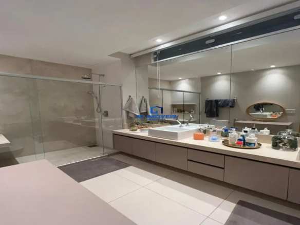 Esta imagem retrata um banheiro amplo, moderno e bem iluminado, com uma paleta de cores predominantemente neutra e sofisticada. Ambiente e Cores: O ambiente geral é clean, espaçoso e elegante. As cores predominantes são tons de bege, branco e cinza claro, criando uma atmosfera calma e relaxante. O teto é branco, e as paredes parecem ser pintadas em um tom de bege muito claro ou off-white, complementando o revestimento. Móveis e Estruturas: Box do Chuveiro: À esquerda, há um espaçoso box de chuveiro com fechamento em vidro temperado transparente e perfis metálicos (provavelmente cromados ou em alumínio). Revestimento Interno: As paredes internas do box são revestidas com grandes placas de cerâmica ou porcelanato em tom bege claro, possivelmente com uma textura sutil ou veios discretos, conferindo um aspecto de pedra natural. Chuveiros: Observam-se dois chuveiros: um de teto, redondo e moderno (tipo "rain shower"), e um chuveiro de mão com barra deslizante, ambos com acabamento metálico. Banco: Dentro do box, há um banco embutido revestido com o mesmo material das paredes, oferecendo conforto e praticidade. Piso do Box: O piso do box parece ser contínuo com o revestimento das paredes, com um ralo linear discreto. Bancada e Pia: Bancada: Uma longa bancada em tom bege claro (possivelmente quartzo, mármore ou material similar) estende-se por grande parte da parede oposta ao box. Cubas: Sobre a bancada, há pelo menos uma cuba de sobrepor retangular, de cor branca e design moderno. Pelo reflexo no espelho e a extensão da bancada, é provável que haja uma segunda cuba, configurando uma bancada dupla. Metais: As torneiras são monocomando, altas, com design contemporâneo e acabamento cromado. Armários: Abaixo da bancada, há um conjunto de armários planejados com portas e gavetas. As frentes dos armários são lisas, sem puxadores aparentes (provavelmente com sistema de abertura por toque ou cava), e apresentam um tom de bege acinzentado ou um laminado amadeirado muito claro e sutil. Espelho e Iluminação: Espelho: Um espelho muito grande cobre toda a parede acima da bancada, do balcão até quase o teto, o que amplia visualmente o espaço e reflete a luz. Iluminação: A iluminação é um destaque, com spots embutidos no teto distribuídos pelo ambiente e uma iluminação linear ou sanca acima do espelho, garantindo que o espaço seja claro e funcional. Piso: O piso do banheiro, fora do box, é revestido com grandes placas de porcelanato em tom branco ou bege muito claro, com acabamento acetinado ou polido. Um tapete retangular cinza está posicionado em frente ao box. Detalhes Adicionais: Toalhas (cinza e bege claro) estão penduradas em suportes. Sobre a bancada, veem-se alguns objetos de higiene pessoal e decoração, como frascos, um pequeno vaso com planta e uma bandeja dourada organizando itens. No primeiro plano, desfocado, há uma superfície que parece ser parte de um móvel ou uma banheira, também em tom claro. No geral, o banheiro transmite uma sensação de luxo discreto, funcionalidade e design contemporâneo, com materiais de boa qualidade e um layout que prioriza o conforto e a amplitude.