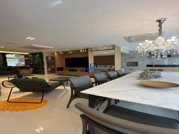 Esta imagem retrata um ambiente interno amplo, moderno e sofisticado, provavelmente uma área de estar e jantar integradas em conceito aberto. Ambiente e Cores: O ambiente é predominantemente claro e luminoso, com um teto branco e um piso de porcelanato brilhante em tom bege claro ou off-white, que reflete a luz e amplia o espaço. A paleta de cores é majoritariamente neutra, com branco, bege, cinza e tons de madeira natural, pontuada por elementos de destaque. O verde vibrante de duas paredes de plantas verticais (jardins verticais) traz vida e um toque natural ao espaço. Há também um tapete redondo em tom laranja vibrante no canto esquerdo, adicionando um ponto de cor quente. Móveis e Detalhes Relevantes: Área de Jantar (Primeiro Plano): Mesa de Jantar: Grande, retangular, com tampo branco que aparenta ser mármore ou quartzo com veios suaves. A base da mesa é escura, possivelmente de madeira ou metal. Cadeiras de Jantar: Design contemporâneo, com estrutura de madeira escura e assento/encosto estofados em tecido ou couro cinza. Lustre: Um suntuoso lustre de cristal pendente sobre a mesa de jantar, com múltiplos braços e pingentes, conferindo um ar clássico e elegante ao espaço moderno. Decoração da Mesa: Minimalista, com um arranjo de plantas (possivelmente suculentas ou tillandsias) e uma fruteira ou objeto decorativo raso em tom dourado/bronze. Área de Estar (Plano Intermediário e Fundo): Chaise Longue e Puff: No lado esquerdo, uma chaise longue de design moderno com estrutura metálica preta fina e estofado em couro ou tecido cinza escuro, acompanhada de um puff no mesmo material. Repousa sobre o tapete laranja. Painel de TV e Rack: Ao fundo, um extenso painel de madeira de tom médio, possivelmente ripado em algumas seções, que ocupa grande parte da parede. Nele está embutida uma grande TV de tela plana. Abaixo da TV, um rack baixo, também em madeira, com compartimentos. Prateleiras e Decoração: Acima e ao lado do painel da TV, prateleiras integradas exibem objetos decorativos como vasos, esculturas pequenas e livros. Sofá: Parcialmente visível mais ao fundo, um sofá grande, aparentemente modular e de cor escura (cinza chumbo ou preto). Paredes Verticais de Plantas: Duas seções de parede são cobertas por jardins verticais com folhagem densa e verde, trazendo um elemento biofílico e textura ao ambiente. Espelho: Uma grande superfície espelhada ao lado de um dos jardins verticais amplia visualmente o espaço e reflete a luz. Cozinha/Área Gourmet (Extremo Fundo à Direita): Uma parede revestida em tom cinza texturizado (possivelmente cimento queimado ou revestimento cerâmico) abriga o que parece ser um forno embutido e talvez uma churrasqueira. Bancada escura (granito preto ou similar) e armários em madeira clara, seguindo o padrão do painel da TV. Iluminação e Outros Detalhes: O teto possui iluminação embutida com múltiplos spots, além do lustre central. Há também unidades de ar condicionado cassete embutidas no teto, indicando climatização central. A sensação geral é de um imóvel de alto padrão, com design de interiores planejado, espaçoso, funcional e elegante, ideal para convívio e entretenimento.