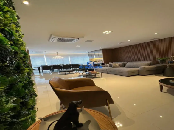 Esta imagem retrata um ambiente interno amplo, moderno e sofisticado, provavelmente uma sala de estar e jantar integradas. Cores e Iluminação: A paleta de cores é predominantemente neutra e elegante. O teto é branco, assim como as paredes visíveis ao fundo, que possuem grandes janelas cobertas por persianas horizontais brancas ou off-white, permitindo a entrada de luz natural difusa. O piso é revestido com grandes lajotas de cor clara, possivelmente bege ou um cinza muito claro, com acabamento brilhante que reflete a luz, ampliando a sensação de espaço. A iluminação artificial é bem distribuída, com múltiplos spots embutidos no teto e uma luminária de teto quadrada com várias lâmpadas. Sobre a mesa de jantar, destaca-se um lustre clássico, com múltiplos braços e elementos que parecem ser cristais. Móveis e Decoração: Sala de Estar: Sofá: Um grande sofá modular em "L" de cor cinza claro domina a área de estar. Possui almofadas decorativas em tons neutros e texturas variadas. Painel de Madeira: A parede à direita do sofá é revestida com um painel ripado de madeira em tom marrom escuro, que adiciona calor e textura ao ambiente. Sobre uma pequena mesa de apoio ao lado do sofá, há um vaso com plantas e objetos decorativos dourados. Mesa de Centro: Há uma mesa de centro mais próxima ao sofá, com estrutura metálica e tampos escuros, possivelmente em madeira ou vidro fumê. Mais à direita, outra mesa de centro redonda com tampo escuro. Poltrona: Em primeiro plano, à esquerda, uma elegante poltrona de design curvo, estofada em couro (ou material similar) na cor caramelo ou marrom claro, com pés finos metálicos dourados. Mesa Lateral/Objeto Decorativo: À frente da poltrona, uma pequena mesa redonda com tampo de madeira clara e uma escultura preta estilizada de um cão (semelhante a um galgo). Parede Verde: À extrema esquerda, uma impressionante parede vertical verde, coberta por folhagens densas (provavelmente um jardim vertical artificial), trazendo um elemento natural e vibrante para o espaço. Sala de Jantar (ao fundo): Mesa de Jantar: Uma longa mesa retangular com tampo escuro (preto ou madeira escura) e várias cadeiras de jantar escuras ao redor. Aparador/Buffet (parcialmente visível): Ao fundo, atrás da sala de jantar e à esquerda do sofá, parece haver um móvel com portas de vidro e iluminação interna, possivelmente um buffet ou uma cristaleira integrada a uma área de bar ou cozinha gourmet. Climatização: Um aparelho de ar condicionado tipo cassete está embutido no teto, indicando preocupação com o conforto térmico. Ambiente: O ambiente transmite uma sensação de luxo contemporâneo, conforto e amplitude. A combinação de materiais como madeira, couro, metal e o toque natural da parede verde cria um espaço equilibrado e acolhedor, ideal para receber e conviver. A organização dos móveis define bem as áreas de estar e jantar, mantendo a fluidez visual do conceito aberto.