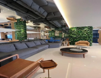 Esta imagem retrata um ambiente de sala de estar amplo e luxuoso, com um design de interiores contemporâneo e sofisticado, característico de um imóvel de alto padrão como o opus urbano areião. O ponto focal imediato é o teto, que é predominantemente espelhado. Este recurso audacioso cria uma ilusão de pé-direito duplicado e reflete todo o ambiente abaixo, incluindo o grande sofá cinza, o piso claro e as paredes. Uma seção do teto é rebaixada e branca, com iluminação embutida indireta que emana um brilho quente e suave. As paredes são um destaque à parte. Grandes painéis de espelho se alternam com exuberantes paredes verdes verticais, que se assemelham a jardins suspensos, trazendo um toque de natureza e frescor para o interior. Ao fundo e à direita, painéis de madeira em tom natural aquecem o ambiente e conferem elegância. O mobiliário é escolhido com esmero: Um sofá secional muito grande, em formato de "L", domina a área de estar. Ele é estofado em um tecido texturizado de cor cinza escuro ou chumbo, com almofadas amplas e confortáveis. Uma mesa de centro oval com tampo escuro (possivelmente vidro preto ou pedra polida) e base de madeira clara está posicionada centralmente. Sobre ela, um objeto decorativo, talvez uma bandeja de pedra clara. Em primeiro plano, uma poltrona de design com estrutura de madeira clara e linhas retas e angulares adiciona um toque de modernidade clássica. Ao lado dela, uma pequena mesa de apoio redonda, também de madeira, com um acessório que parece ser uma bandeja de couro marrom. Mais ao fundo, próxima a uma das paredes verdes, há uma poltrona individual em couro caramelo ou bege, com pés metálicos finos, conferindo leveza. O piso é revestido com grandes placas de porcelanato em tom claro, possivelmente branco ou off-white, o que contribui para a sensação de amplitude e luminosidade, além de refletir a luz natural e artificial. A iluminação é bem planejada, combinando a luz indireta do teto rebaixado com outros pontos de luz que não são diretamente visíveis, mas cujos reflexos são notados nos espelhos, criando uma atmosfera acolhedora e elegante. No geral, o ambiente transmite uma sensação de sofisticação, modernidade e conforto. A paleta de cores é neutra e elegante, com o cinza, branco, tons de madeira e o verde vibrante das plantas criando um contraste harmonioso. A utilização extensiva de espelhos e o pé-direito generoso (ampliado visualmente) reforçam a sensação de espaço e luxo.