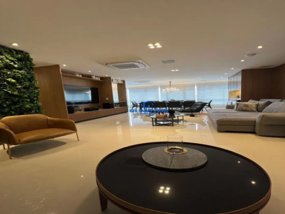 Esta imagem retrata um ambiente interno amplo, moderno e sofisticado, provavelmente uma sala de estar e jantar integrada. Cores e Materiais: A paleta de cores é predominantemente neutra e elegante. O piso é de um material claro, possivelmente porcelanato polido em tom bege ou off-white, com alto brilho, refletindo a iluminação e os móveis. As paredes laterais e o teto são brancos. Destacam-se painéis de madeira em tom claro a médio, revestindo parte das paredes, como a que abriga a TV e a parede ao fundo do sofá, conferindo aconchego e textura. Há uma grande parede vertical coberta por um jardim vertical (ou painel de plantas artificiais) de um verde vibrante e denso, trazendo um elemento natural e um ponto focal de cor. O mobiliário combina tons de marrom (couro caramelo da poltrona), cinza (sofá) e preto (mesas de centro e de jantar). Detalhes metálicos em dourado ou latão estão presentes nos pés da poltrona e em um adorno na mesa de centro. Ambiente e Iluminação: O ambiente é espaçoso e bem iluminado. O teto possui diversas luminárias embutidas (spots de LED), algumas em conjuntos de quatro, distribuindo a luz de forma uniforme. Um grande lustre de cristal ou vidro com múltiplas lâmpadas pende sobre a mesa de jantar ao fundo, adicionando um toque de luxo clássico. Grandes janelas ao fundo, cobertas por persianas horizontais claras, permitem a entrada de luz natural controlada. A sensação geral é de um espaço luxuoso, contemporâneo e bem planejado, com uma atmosfera acolhedora apesar da modernidade. Há também um sistema de ar condicionado central embutido no teto. Móveis: Primeiro Plano: Uma mesa de centro redonda com tampo preto brilhante e borda amadeirada. Sobre ela, um elemento decorativo circular em pedra escura (ardósia ou similar) e um vaso cilíndrico de vidro com detalhe metálico dourado. Sala de Estar (Esquerda): Uma poltrona ou pequeno sofá de dois lugares em couro caramelo, com design curvilíneo e pés finos metálicos dourados, posicionada em frente à parede verde. Um longo rack de madeira clara com detalhes ripados na parte inferior, sobre o qual está uma grande TV de tela plana. Prateleiras acima da TV exibem objetos decorativos discretos e caixas de som. Sala de Estar (Direita): Um grande sofá modular em "L" estofado em tecido cinza claro, com várias almofadas, algumas lisas e outras com padrões geométricos. Sala de Jantar (Fundo): Uma grande mesa de jantar, aparentemente preta ou de tom muito escuro, rodeada por múltiplas cadeiras de design moderno, também escuras. Sobre a mesa, alguns objetos decorativos, possivelmente em tons de azul. Próximo à área de jantar, uma chaise longue ou poltrona reclinável preta, acompanhada por uma pequena mesa de apoio preta. Detalhes Adicionais: Ao fundo, próximo à sala de jantar e ao sofá, parece haver uma estante ou cristaleira com prateleiras de vidro e fundo espelhado, refletindo a luz. O design de interiores privilegia linhas retas e formas geométricas, com um bom equilíbrio entre materiais naturais (madeira, plantas) e elementos modernos e sofisticados (vidro, metal, superfícies polidas).