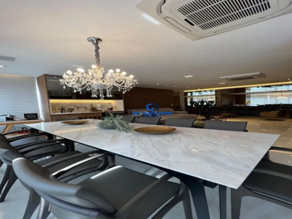 Esta imagem retrata um ambiente interno amplo, moderno e sofisticado, provavelmente uma sala de jantar integrada a uma sala de estar. Sala de Jantar (Primeiro Plano): O foco principal é uma longa e imponente mesa de jantar retangular com tampo branco, possivelmente de mármore ou quartzo com veios suaves em tons de cinza, conferindo um aspecto luxuoso e limpo. A base da mesa é escura, provavelmente preta, com pernas robustas de design contemporâneo. Ao redor da mesa, há várias cadeiras com braços, estofadas em um material que parece couro ou courino de cor cinza escuro ou preta, com estruturas também escuras, harmonizando com a base da mesa. O design das cadeiras é moderno e confortável. Sobre a mesa, como decoração, há dois recipientes baixos e largos de tom dourado ou bronze, e um arranjo central com plantas verdes, possivelmente suculentas, em um vaso baixo de vidro. Suspenso sobre a mesa, destaca-se um lustre de cristal clássico e opulento, com múltiplos braços, lâmpadas que imitam velas e pingentes de cristal, criando um contraste interessante entre o clássico e o moderno e adicionando um toque de glamour ao ambiente. Ambiente Geral e Fundo: O piso é claro, de grandes placas, possivelmente porcelanato polido, que reflete a luz e contribui para a sensação de amplitude. O teto é branco e conta com iluminação embutida (spots) e uma unidade de ar condicionado central do tipo cassete. À esquerda da sala de jantar, há uma grande janela coberta por persianas horizontais brancas, permitindo a entrada de luz natural difusa. Abaixo dela, parece haver uma bancada ou mesa de trabalho com alguns equipamentos, como uma impressora. Próximo a esta área, há um móvel aparador ou buffet embutido, com nichos iluminados internamente e prateleiras, combinando tons de madeira clara e elementos escuros ou espelhados. Ao fundo, vislumbra-se a sala de estar. Ela é composta por um sofá grande, seccional, em tom claro (possivelmente cinza claro ou bege). Atrás do sofá, uma parede é revestida com um painel de madeira ripada em tom escuro, adicionando textura e aconchego. Há também uma poltrona em tom caramelo ou marrom claro. Plantas ornamentais e grandes painéis de espelho ao fundo ampliam visualmente o espaço e refletem a luz, intensificando a luminosidade. Cores e Atmosfera: A paleta de cores é predominantemente neutra e sofisticada: branco, cinza (em várias tonalidades), preto e tons de madeira. Os detalhes em dourado/bronze do lustre e dos objetos na mesa, e o verde das plantas, adicionam pontos de cor e interesse visual. A atmosfera geral é de luxo contemporâneo, elegância e conforto. O espaço é bem iluminado, tanto por luz natural quanto artificial, e parece ser funcional e bem planejado para convívio e entretenimento.