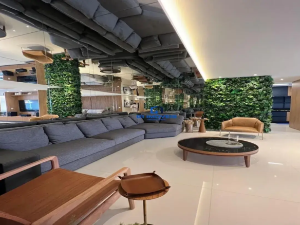 Esta imagem retrata um ambiente de sala de estar amplo e luxuoso, com um design de interiores contemporâneo e sofisticado, característico de um imóvel de alto padrão como o opus urbano areião. O ponto focal imediato é o teto, que é predominantemente espelhado. Este recurso audacioso cria uma ilusão de pé-direito duplicado e reflete todo o ambiente abaixo, incluindo o grande sofá cinza, o piso claro e as paredes. Uma seção do teto é rebaixada e branca, com iluminação embutida indireta que emana um brilho quente e suave. As paredes são um destaque à parte. Grandes painéis de espelho se alternam com exuberantes paredes verdes verticais, que se assemelham a jardins suspensos, trazendo um toque de natureza e frescor para o interior. Ao fundo e à direita, painéis de madeira em tom natural aquecem o ambiente e conferem elegância. O mobiliário é escolhido com esmero: Um sofá secional muito grande, em formato de "L", domina a área de estar. Ele é estofado em um tecido texturizado de cor cinza escuro ou chumbo, com almofadas amplas e confortáveis. Uma mesa de centro oval com tampo escuro (possivelmente vidro preto ou pedra polida) e base de madeira clara está posicionada centralmente. Sobre ela, um objeto decorativo, talvez uma bandeja de pedra clara. Em primeiro plano, uma poltrona de design com estrutura de madeira clara e linhas retas e angulares adiciona um toque de modernidade clássica. Ao lado dela, uma pequena mesa de apoio redonda, também de madeira, com um acessório que parece ser uma bandeja de couro marrom. Mais ao fundo, próxima a uma das paredes verdes, há uma poltrona individual em couro caramelo ou bege, com pés metálicos finos, conferindo leveza. O piso é revestido com grandes placas de porcelanato em tom claro, possivelmente branco ou off-white, o que contribui para a sensação de amplitude e luminosidade, além de refletir a luz natural e artificial. A iluminação é bem planejada, combinando a luz indireta do teto rebaixado com outros pontos de luz que não são diretamente visíveis, mas cujos reflexos são notados nos espelhos, criando uma atmosfera acolhedora e elegante. No geral, o ambiente transmite uma sensação de sofisticação, modernidade e conforto. A paleta de cores é neutra e elegante, com o cinza, branco, tons de madeira e o verde vibrante das plantas criando um contraste harmonioso. A utilização extensiva de espelhos e o pé-direito generoso (ampliado visualmente) reforçam a sensação de espaço e luxo.