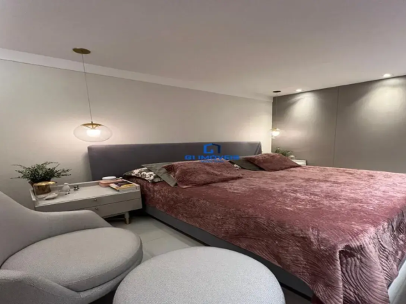 Esta imagem retrata um quarto de casal com um design moderno e elegante, transmitindo uma atmosfera acolhedora e sofisticada. Cores e Materiais: A paleta de cores é predominantemente neutra e suave, com toques de cor que adicionam calor e profundidade. Paredes: A parede principal, à esquerda, é pintada em um tom de cinza claro ou off-white. A parede de destaque, atrás da cabeceira da cama, parece ser revestida com painéis de madeira ou um papel de parede texturizado em um tom de cinza mais escuro e quente, criando um contraste sutil e elegante. Teto: Branco, com sancas discretas e iluminação embutida. Piso: Claro, possivelmente de porcelanato ou vinílico em tom neutro, contribuindo para a sensação de amplitude. Roupa de Cama: A colcha é o destaque de cor, em um tom de rosa antigo ou marsala, com uma textura aveludada e um padrão sutil em relevo, como ondas ou drapeados. Os travesseiros combinam essa cor com outros em tons de cinza escuro e um com estampa (possivelmente floral ou abstrata em tons neutros), adicionando camadas e interesse visual. Móveis: Predominantemente em tons de cinza. Detalhes Metálicos: Toques de dourado/latão nos pendentes e nos pés da mesa de cabeceira conferem um ar de sofisticação. Móveis e Decoração: Cama: Cama de casal com uma cabeceira alta e estofada em tecido cinza escuro, possivelmente veludo ou um tecido similar, que se estende para além da largura da cama. Mesas de Cabeceira: A mesa de cabeceira visível à esquerda é de design contemporâneo, na cor cinza claro, com gavetas e pés finos, possivelmente com pontas metálicas douradas. Sobre ela, há um pequeno vaso com uma planta, um livro, um objeto decorativo pequeno (como um pássaro) e um pote com tampa rosa/coral. Poltrona e Pufe: Em primeiro plano, há uma poltrona de design moderno, com formas arredondadas e estofamento em tecido texturizado cinza claro (talvez bouclé). À frente dela, um pufe redondo, aparentemente com o mesmo tecido, completa um pequeno recanto de estar ou leitura. Iluminação: A iluminação é um ponto forte do ambiente. Pendentes: Dois pendentes com design moderno, compostos por um globo de vidro transparente ou fosco e detalhes em metal dourado, estão posicionados sobre as mesas de cabeceira (um de cada lado da cama). Spots Embutidos: No teto, há spots de luz direcionados para a parede de destaque atrás da cama, realçando sua textura e cor. Objetos Decorativos: Além dos itens na mesa de cabeceira, a decoração é minimalista, focando na qualidade dos materiais e no design dos móveis. Ambiente: O ambiente geral é de um quarto luxuoso, confortável e bem planejado. A iluminação quente e as texturas macias (veludo, tecido da poltrona) criam uma sensação de aconchego e intimidade, ideal para descanso. O uso de cores neutras com o ponto de cor na roupa de cama torna o espaço elegante e contemporâneo, sem ser frio. A disposição dos móveis sugere um espaço funcional e bem aproveitado.