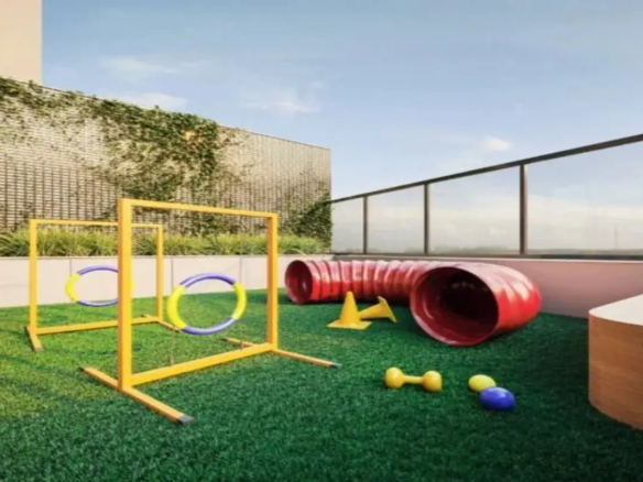 A imagem mostra um espaço pet (playground para animais de estimação) localizado numa área externa, provavelmente o terraço ou rooftop de um edifício residencial moderno. O ambiente é aberto e bem iluminado pela luz natural do dia, sob um céu azul claro com poucas nuvens. Detalhes do Imóvel e Ambiente: Piso: O chão é coberto por grama sintética de um verde intenso e vibrante, proporcionando uma superfície macia e adequada para os animais. Paredes e Limites: Ao fundo, há uma parede alta com uma textura que parece ser uma treliça ou grade bege claro, parcialmente coberta por vegetação trepadeira verde, conferindo um toque natural ao espaço. Acima desta parede, vê-se a lateral de um prédio mais alto, de cor clara. À direita, a área é delimitada por um guarda-corpo moderno de vidro transparente com postes e corrimão em metal escuro (provavelmente preto ou cinza chumbo), garantindo segurança sem obstruir a vista. Iluminação e Atmosfera: A luz é natural e abundante, criando um ambiente ensolarado e convidativo. A atmosfera geral é limpa, organizada e projetada especificamente para a recreação de animais. Móveis e Equipamentos (Foco Pet): O espaço está equipado com vários itens de agility para cães: Obstáculos de Salto (Aros): Dois conjuntos de obstáculos de salto com estruturas metálicas pintadas de amarelo vibrante. Cada estrutura sustenta um aro circular para o cão pular através. Os aros são coloridos em segmentos alternados de azul e amarelo. Túnel: Um túnel flexível, sanfonado, de cor vermelho vivo, posicionado sobre a grama. É um equipamento clássico para cães correrem por dentro. Cones: Dois pequenos cones de sinalização na cor amarela, geralmente usados para treinos de slalom ou demarcação. Plataforma/Banco: À direita, próximo ao guarda-corpo, há uma estrutura baixa e curva, que parece ser um pequeno banco ou plataforma, com acabamento que imita madeira clara. Pode servir como obstáculo baixo ou local de descanso. Brinquedos: Espalhados pela grama, há alguns brinquedos para cães: um haltere pequeno de borracha amarela, uma bola amarela e uma bola azul. Cores: A paleta de cores é vibrante e alegre, dominada pelo verde da grama, o azul do céu, e os tons primários brilhantes (amarelo e vermelho) dos equipamentos, contrastando com os tons neutros da parede de fundo e do guarda-corpo. Em resumo, a imagem retrata uma área de lazer externa moderna, segura e funcional, projetada como um diferencial do imóvel, oferecendo um espaço dedicado e equipado para a diversão e exercício de animais de estimação.
