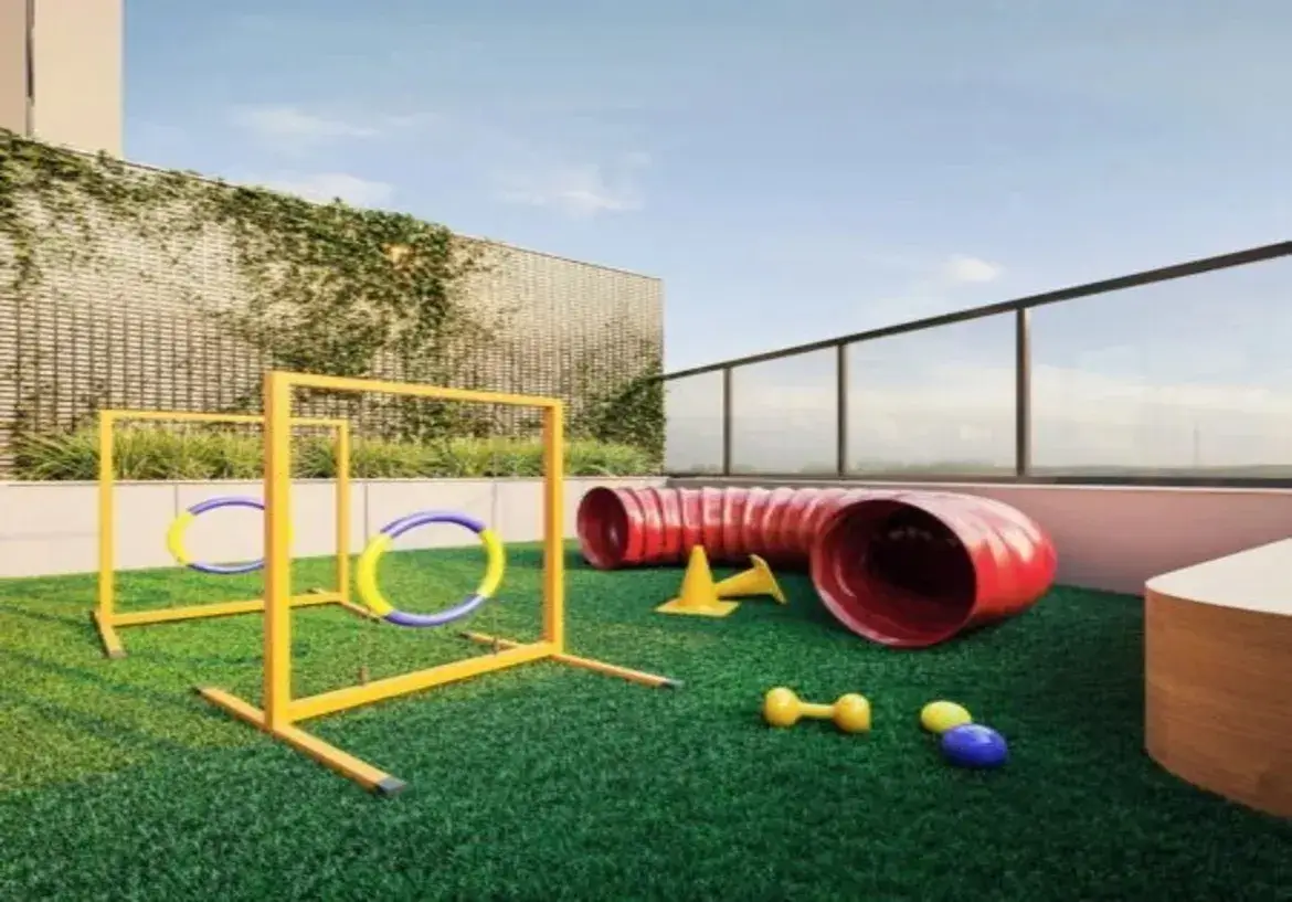 A imagem mostra um espaço pet (playground para animais de estimação) localizado numa área externa, provavelmente o terraço ou rooftop de um edifício residencial moderno. O ambiente é aberto e bem iluminado pela luz natural do dia, sob um céu azul claro com poucas nuvens. Detalhes do Imóvel e Ambiente: Piso: O chão é coberto por grama sintética de um verde intenso e vibrante, proporcionando uma superfície macia e adequada para os animais. Paredes e Limites: Ao fundo, há uma parede alta com uma textura que parece ser uma treliça ou grade bege claro, parcialmente coberta por vegetação trepadeira verde, conferindo um toque natural ao espaço. Acima desta parede, vê-se a lateral de um prédio mais alto, de cor clara. À direita, a área é delimitada por um guarda-corpo moderno de vidro transparente com postes e corrimão em metal escuro (provavelmente preto ou cinza chumbo), garantindo segurança sem obstruir a vista. Iluminação e Atmosfera: A luz é natural e abundante, criando um ambiente ensolarado e convidativo. A atmosfera geral é limpa, organizada e projetada especificamente para a recreação de animais. Móveis e Equipamentos (Foco Pet): O espaço está equipado com vários itens de agility para cães: Obstáculos de Salto (Aros): Dois conjuntos de obstáculos de salto com estruturas metálicas pintadas de amarelo vibrante. Cada estrutura sustenta um aro circular para o cão pular através. Os aros são coloridos em segmentos alternados de azul e amarelo. Túnel: Um túnel flexível, sanfonado, de cor vermelho vivo, posicionado sobre a grama. É um equipamento clássico para cães correrem por dentro. Cones: Dois pequenos cones de sinalização na cor amarela, geralmente usados para treinos de slalom ou demarcação. Plataforma/Banco: À direita, próximo ao guarda-corpo, há uma estrutura baixa e curva, que parece ser um pequeno banco ou plataforma, com acabamento que imita madeira clara. Pode servir como obstáculo baixo ou local de descanso. Brinquedos: Espalhados pela grama, há alguns brinquedos para cães: um haltere pequeno de borracha amarela, uma bola amarela e uma bola azul. Cores: A paleta de cores é vibrante e alegre, dominada pelo verde da grama, o azul do céu, e os tons primários brilhantes (amarelo e vermelho) dos equipamentos, contrastando com os tons neutros da parede de fundo e do guarda-corpo. Em resumo, a imagem retrata uma área de lazer externa moderna, segura e funcional, projetada como um diferencial do imóvel, oferecendo um espaço dedicado e equipado para a diversão e exercício de animais de estimação.