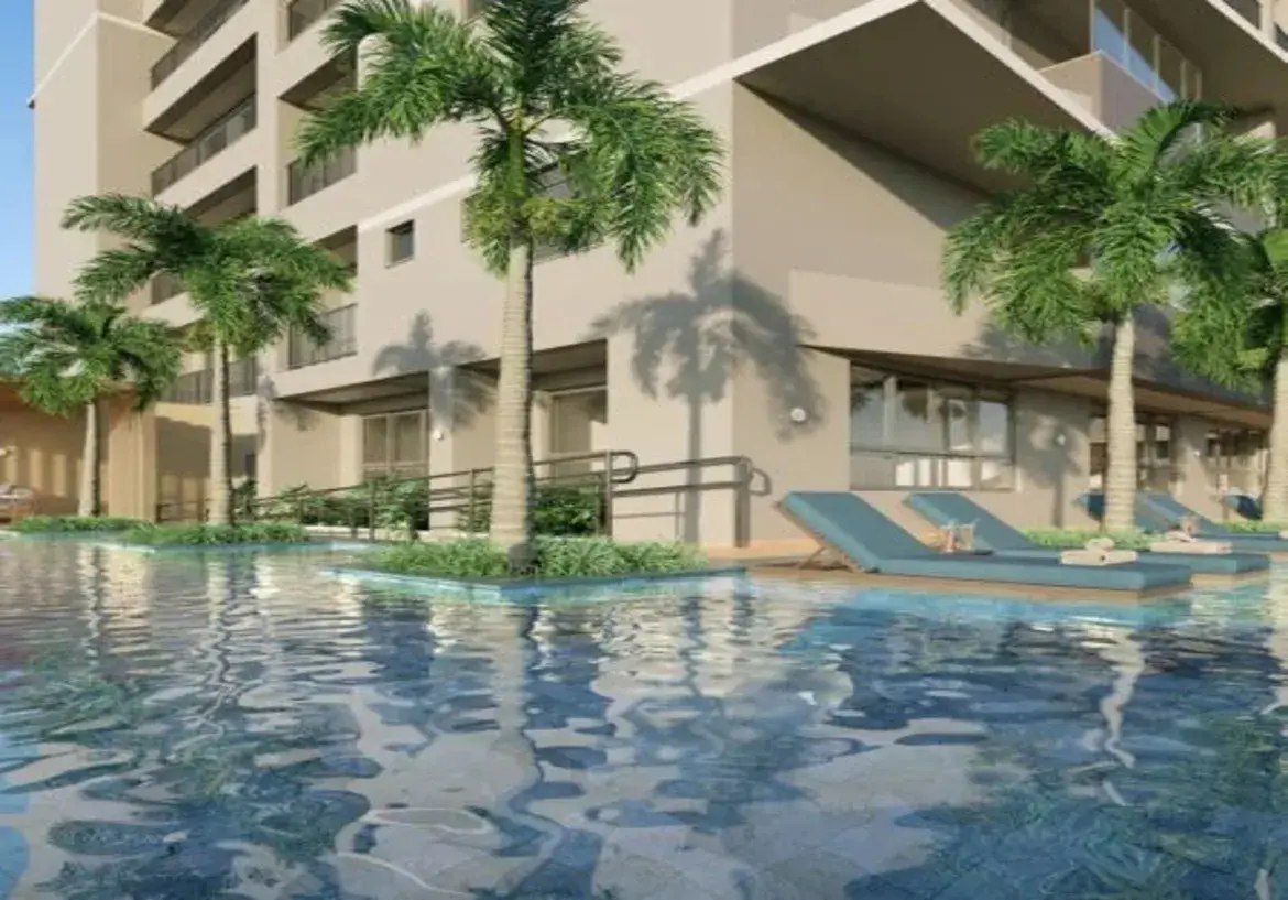 A imagem apresenta uma vista ao nível da água da área da piscina de um edifício residencial moderno. Imóvel (Edifício): Estrutura: Trata-se de um prédio de múltiplos andares com arquitetura contemporânea. Possui linhas retas, varandas com guarda-corpos (aparentemente de metal ou vidro) e janelas de diferentes tamanhos. Cores: A fachada do edifício é predominantemente pintada em um tom neutro, como bege claro ou um branco "off-white". Há áreas de sombra sob as varandas e beirais que criam contraste. Detalhes: No térreo, junto à piscina, percebe-se uma área com grandes janelas, possivelmente uma área comum ou apartamentos com acesso direto. Nota-se também uma rampa de acesso com corrimão escuro (provavelmente metal), indicando acessibilidade. Ambiente: Atmosfera: O ambiente geral é de lazer, relaxamento e sofisticação, típico de uma área comum de condomínio ou resort. A iluminação sugere um dia claro e ensolarado, com sombras bem definidas projetadas pelas palmeiras na fachada do prédio. Paisagismo: A área é adornada com várias palmeiras altas e esguias, com troncos claros e folhagem verde vibrante, que conferem um ar tropical ao local. Há também vegetação mais baixa (arbustos ou folhagens) plantada em canteiros na borda da piscina, integrando a natureza ao espaço construído. Piscina: A piscina ocupa o primeiro plano. A água é azul, límpida e apresenta leves ondulações na superfície, refletindo o céu e o entorno. O revestimento interno parece ser de pastilhas ou azulejos em tons de azul e talvez cinza, visíveis através da água. A perspectiva baixa, ao nível da água, cria uma sensação de imersão. Móveis: Espreguiçadeiras: Dispostas sobre um deck (que parece ser de madeira clara ou material composto) à beira da piscina, há várias espreguiçadeiras. Cor e Material: Elas se destacam pela cor vibrante de seus colchonetes, um azul-petróleo ou azul-turquesa. A estrutura das espreguiçadeiras parece ser de material claro, possivelmente alumínio ou madeira tratada. Detalhes: Algumas espreguiçadeiras possuem toalhas brancas dobradas sobre elas, reforçando a sensação de conforto e prontidão para uso. Em resumo, a imagem mostra uma área de piscina convidativa e bem projetada, integrada a um edifício residencial de cores neutras. O contraste entre o azul da água e das espreguiçadeiras, o verde das palmeiras e o bege do prédio cria uma composição visual harmoniosa e atraente, transmitindo uma sensação de tranquilidade e lazer.