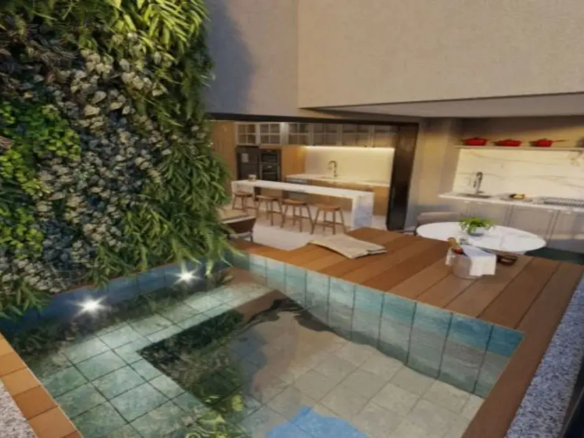 A imagem mostra um ambiente residencial interno/externo integrado, com um design moderno e sofisticado. O espaço parece ser uma área de lazer ou gourmet de um apartamento de alto padrão ou cobertura. Piscina/Jacuzzi: Em primeiro plano e à esquerda, há uma piscina ou jacuzzi em formato de "L". Ela é revestida internamente com pastilhas pequenas, que aparentam ser de um tom azul-esverdeado claro ou turquesa, conferindo uma cor vibrante à água cristalina. A borda da piscina possui iluminação embutida (spots de luz) que clareiam a água. A margem externa da piscina é feita com ladrilhos de cor terracota. Parede Verde Vertical: Dominando a parede lateral esquerda, adjacente à piscina, existe um jardim vertical exuberante. É composto por uma densa variedade de folhagens verdes, incluindo samambaias e outras plantas, em diferentes tonalidades e texturas. Esta parede viva traz um forte elemento natural para o ambiente, contrastando com os materiais mais sóbrios e adicionando frescor. Deck de Madeira: Ao lado da piscina, estende-se um deck de madeira ripada, em um tom marrom médio, quente. Ele funciona como uma área de estar e transição entre a piscina e o espaço gourmet/cozinha. Sobre o deck, há uma toalha ou manta bege dobrada. Área de Estar/Refeição: No lado direito do deck, há uma pequena mesa redonda com tampo branco (possivelmente mármore) e um vaso com uma planta verde no centro. Ao lado da mesa, um balde de gelo prateado com uma garrafa (provavelmente espumante ou vinho branco) e um guardanapo branco sugerem um espaço preparado para relaxamento e celebração. Cozinha Integrada (ao fundo, à esquerda): Mais ao fundo, visível através de uma abertura ampla, encontra-se uma cozinha moderna. Ela possui uma ilha central com tampo branco (mármore ou quartzo) que serve como balcão para refeições rápidas, equipada com quatro banquetas altas de madeira clara com design simples. Os armários da cozinha combinam tons de madeira clara ou bege na base e possivelmente armários superiores em cor clara ou com portas de vidro. A iluminação sob os armários superiores destaca o backsplash claro. Um refrigerador escuro (preto ou inox) está embutido. O piso da cozinha parece ser de porcelanato em tom neutro claro (bege ou cinza). Espaço Gourmet (ao fundo, à direita): Complementando a cozinha, à direita, há outra bancada de apoio, também com tampo branco de aparência marmorizada e armários inferiores brancos. Um backsplash do mesmo material da bancada sobe pela parede. Uma pia está instalada nesta bancada. Acima, uma prateleira branca suspensa exibe objetos decorativos, incluindo três panelas ou caçarolas vermelhas que adicionam um ponto de cor ao espaço. Ambiente e Cores: A atmosfera geral é de luxo, conforto e modernidade. A paleta de cores é predominantemente neutra, com muito branco, bege, cinza e tons de madeira natural. O verde vibrante do jardim vertical e o azul-esverdeado da piscina criam pontos focais de cor e trazem vida ao ambiente. Os detalhes em vermelho (panelas) e prata (balde de gelo) são sutis, mas adicionam elegância. A iluminação parece ser bem planejada, combinando luz natural (implícita) com luz artificial (spots na piscina, luz sob armários), criando um ambiente acolhedor e convidativo, tanto para o dia quanto para a noite. A integração dos espaços (piscina, deck, cozinha, área gourmet) sugere um local projetado para o convívio social e o lazer.