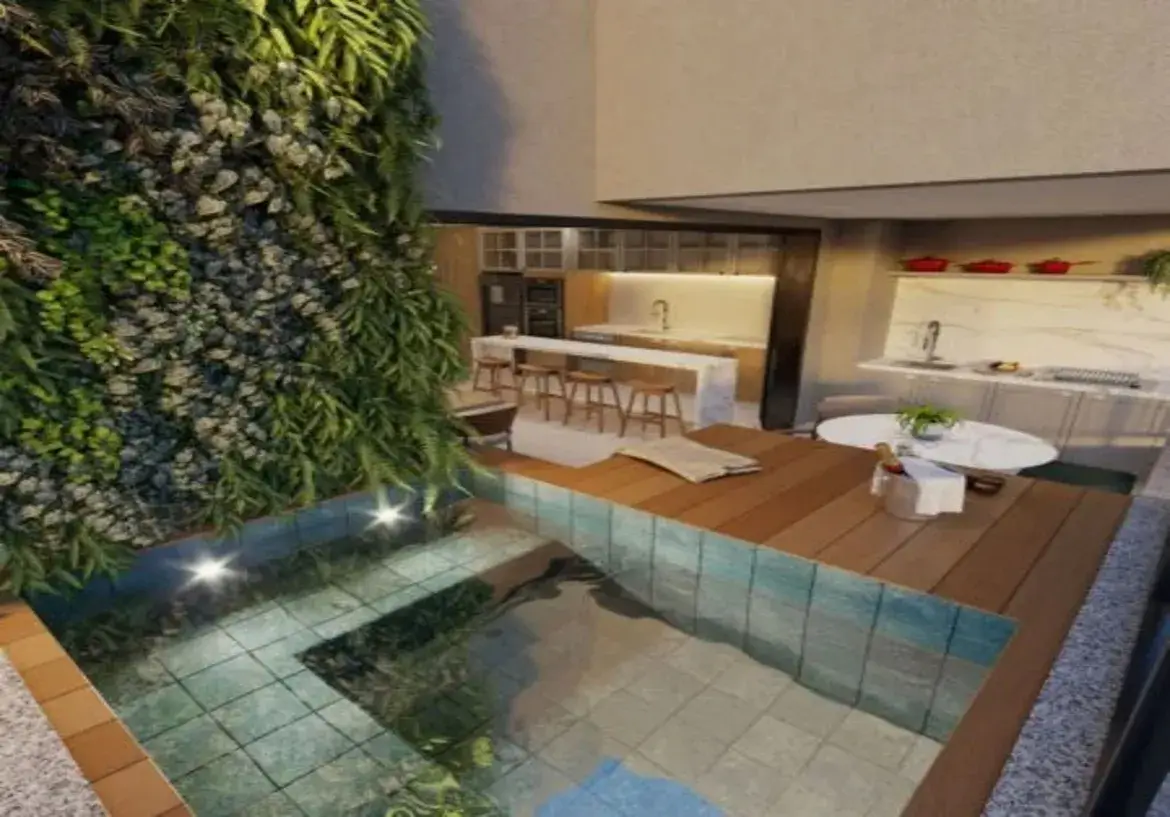 A imagem mostra um ambiente residencial interno/externo integrado, com um design moderno e sofisticado. O espaço parece ser uma área de lazer ou gourmet de um apartamento de alto padrão ou cobertura. Piscina/Jacuzzi: Em primeiro plano e à esquerda, há uma piscina ou jacuzzi em formato de "L". Ela é revestida internamente com pastilhas pequenas, que aparentam ser de um tom azul-esverdeado claro ou turquesa, conferindo uma cor vibrante à água cristalina. A borda da piscina possui iluminação embutida (spots de luz) que clareiam a água. A margem externa da piscina é feita com ladrilhos de cor terracota. Parede Verde Vertical: Dominando a parede lateral esquerda, adjacente à piscina, existe um jardim vertical exuberante. É composto por uma densa variedade de folhagens verdes, incluindo samambaias e outras plantas, em diferentes tonalidades e texturas. Esta parede viva traz um forte elemento natural para o ambiente, contrastando com os materiais mais sóbrios e adicionando frescor. Deck de Madeira: Ao lado da piscina, estende-se um deck de madeira ripada, em um tom marrom médio, quente. Ele funciona como uma área de estar e transição entre a piscina e o espaço gourmet/cozinha. Sobre o deck, há uma toalha ou manta bege dobrada. Área de Estar/Refeição: No lado direito do deck, há uma pequena mesa redonda com tampo branco (possivelmente mármore) e um vaso com uma planta verde no centro. Ao lado da mesa, um balde de gelo prateado com uma garrafa (provavelmente espumante ou vinho branco) e um guardanapo branco sugerem um espaço preparado para relaxamento e celebração. Cozinha Integrada (ao fundo, à esquerda): Mais ao fundo, visível através de uma abertura ampla, encontra-se uma cozinha moderna. Ela possui uma ilha central com tampo branco (mármore ou quartzo) que serve como balcão para refeições rápidas, equipada com quatro banquetas altas de madeira clara com design simples. Os armários da cozinha combinam tons de madeira clara ou bege na base e possivelmente armários superiores em cor clara ou com portas de vidro. A iluminação sob os armários superiores destaca o backsplash claro. Um refrigerador escuro (preto ou inox) está embutido. O piso da cozinha parece ser de porcelanato em tom neutro claro (bege ou cinza). Espaço Gourmet (ao fundo, à direita): Complementando a cozinha, à direita, há outra bancada de apoio, também com tampo branco de aparência marmorizada e armários inferiores brancos. Um backsplash do mesmo material da bancada sobe pela parede. Uma pia está instalada nesta bancada. Acima, uma prateleira branca suspensa exibe objetos decorativos, incluindo três panelas ou caçarolas vermelhas que adicionam um ponto de cor ao espaço. Ambiente e Cores: A atmosfera geral é de luxo, conforto e modernidade. A paleta de cores é predominantemente neutra, com muito branco, bege, cinza e tons de madeira natural. O verde vibrante do jardim vertical e o azul-esverdeado da piscina criam pontos focais de cor e trazem vida ao ambiente. Os detalhes em vermelho (panelas) e prata (balde de gelo) são sutis, mas adicionam elegância. A iluminação parece ser bem planejada, combinando luz natural (implícita) com luz artificial (spots na piscina, luz sob armários), criando um ambiente acolhedor e convidativo, tanto para o dia quanto para a noite. A integração dos espaços (piscina, deck, cozinha, área gourmet) sugere um local projetado para o convívio social e o lazer.
