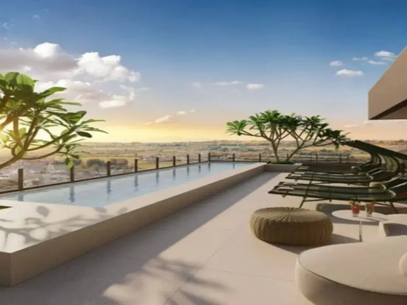 A imagem retrata uma luxuosa área de lazer na cobertura de um imóvel, provavelmente um prédio residencial de alto padrão ou um hotel. O ponto focal é uma piscina de borda infinita (ou com uma borda elevada que cria um efeito semelhante), longa e retangular, com água calma e cristalina que reflete o céu. A estrutura da piscina é robusta, feita de um material claro, possivelmente concreto ou revestimento em tons de bege ou branco-gelo. Ao longo da borda da piscina que dá para a vista, há um guarda-corpo de vidro transparente, garantindo segurança sem obstruir a paisagem. O ambiente geral é moderno, sofisticado e relaxante, acentuado pela iluminação de um pôr do sol ou nascer do sol. O céu exibe um azul vibrante na parte superior, salpicado por nuvens brancas e fofas, enquanto próximo ao horizonte predominam tons quentes de amarelo e laranja, onde o sol brilha intensamente, criando um contraluz em uma das árvores. O piso da área externa é revestido com ladrilhos grandes ou cimento queimado em um tom neutro claro, como bege ou off-white, harmonizando com a borda da piscina e contribuindo para a sensação de amplitude e limpeza. Quanto aos móveis, dispostos sobre o deck, temos: Uma fileira de espreguiçadeiras de design contemporâneo, com estrutura fina (aparentemente metálica) e assento em trama ou tela de cor escura (verde-musgo escuro, marrom ou preto). Estão posicionadas para aproveitar a vista e o sol. Um pufe redondo de trama natural (como rattan ou similar) em tom bege/castanho claro, servindo como assento extra ou apoio. Ao lado do pufe, uma pequena mesa de apoio redonda e branca, sobre a qual repousam dois copos com bebidas coloridas (avermelhadas), sugerindo um momento de descontração. Em primeiro plano, parcialmente visível, há um grande sofá ou daybed redondo, estofado em tecido claro (branco ou creme), de aparência muito confortável. A vegetação inclui algumas árvores de porte médio com folhagem verdejante (lembrando Plumerias/Jasmim-manga), estrategicamente posicionadas para adicionar um toque natural e de frescor ao ambiente, sem bloquear a vista panorâmica da cidade e da paisagem mais distante que se estende até o horizonte. A paleta de cores é predominantemente neutra e clara (bege, branco, cinza claro) na estrutura e no piso, contrastando com o azul da piscina e do céu, os tons quentes do sol, o verde das plantas e o tom escuro das espreguiçadeiras. Em resumo, a imagem descreve um espaço de lazer exclusivo e elegante, projetado para oferecer conforto, relaxamento e uma vista deslumbrante, ideal para desfrutar de momentos ao ar livre em grande estilo.