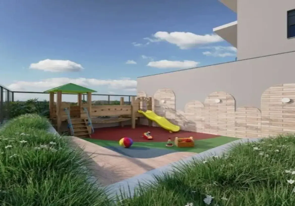 Descrição Geral: A imagem retrata um parquinho infantil (playground) ao ar livre, localizado provavelmente em uma área comum de um condomínio ou prédio residencial, possivelmente um terraço ou área externa elevada, dado a presença de uma cerca e a vista do céu. O ambiente geral é ensolarado, moderno e projetado para ser seguro e divertido para crianças. Detalhes do Imóvel e Ambiente: Piso: A área principal do playground possui um piso emborrachado de segurança, dividido em duas cores principais: uma seção maior em vermelho-terracota e uma seção menor em verde-escuro. Este tipo de piso é ideal para absorver impactos. Ao redor dessa área colorida, há um caminho ou borda em um tom de concreto claro ou bege. Paredes e Estrutura: À direita, uma parede alta e lisa na cor cinza-claro ou off-white domina parte da cena. Na base desta parede, há um revestimento decorativo mais baixo, feito de tijolinhos ou placas em tons de bege-claro, com um design ondulado no topo, lembrando ameias de castelo simplificadas. Pequenas luminárias redondas estão instaladas sobre essa parte decorativa. Ao fundo, vê-se a estrutura de um prédio na mesma cor cinza-claro. Cerca e Paisagismo: Ao fundo e à esquerda, uma cerca de segurança preta com tela delimita o espaço. Atrás da cerca, há vegetação (arbustos verdes), sugerindo um limite da propriedade ou a borda de um terraço. Em primeiro plano e ladeando o caminho de acesso ao parquinho, há canteiros elevados ou floreiras com grama verde vibrante e pequenas flores brancas, que trazem um elemento natural e suavizam o design. Céu e Iluminação: O céu está azul claro com algumas nuvens brancas e fofas, indicando um dia de tempo bom e ensolarado. A iluminação geral é natural e brilhante. Móveis e Equipamentos (Brinquedos): Estrutura Principal do Playground: O equipamento central é uma estrutura de playground multifuncional feita predominantemente de madeira clara (pinus ou similar). Possui: Uma pequena torre com telhado verde. Escadas de acesso com degraus de madeira e detalhes laterais azuis. Uma seção que parece ser uma pequena parede de escalada com agarras coloridas. Um escorregador amarelo vibrante, de plástico, com formato levemente ondulado, que desce da estrutura até o piso emborrachado verde. Brinquedos Soltos: Sobre o piso emborrachado verde, há alguns brinquedos infantis: Uma bola colorida (listras vermelhas, amarelas, azuis e verdes). Um pequeno veículo de empurrar ou montar, vermelho. Uma caixa de madeira clara contendo alguns objetos (possivelmente blocos ou frutas de brinquedo). Um brinquedo de empilhar próximo à caixa. Cores Predominantes: A paleta de cores combina tons naturais (madeira clara, bege dos tijolos, verde da grama) com cores vibrantes e primárias (amarelo do escorregador, vermelho e verde do piso, cores da bola e brinquedos) e tons neutros (cinza-claro da parede e do prédio, preto da cerca). O azul do céu complementa a cena. Em resumo, a imagem mostra um espaço de lazer infantil bem equipado, seguro e esteticamente agradável, com uma mistura equilibrada de materiais naturais, cores vibrantes e design moderno, inserido em um contexto residencial.