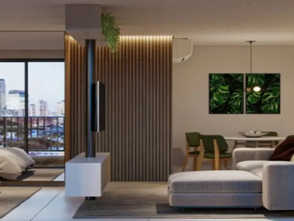 A imagem mostra o interior de um apartamento moderno, com um design de conceito aberto que integra sala de estar, sala de jantar e, parcialmente, o quarto. O ambiente geral é contemporâneo, funcional e aconchegante, com uma paleta de cores predominantemente neutra, pontuada por toques de cor e texturas naturais. Layout e Estrutura: O espaço é dividido de forma inteligente. Uma estrutura central funciona como divisor de ambientes e ponto focal: um pilar metálico escuro que vai do chão ao teto sustenta uma TV de tela plana. Abaixo da TV, um móvel baixo (rack/console) branco e flutuante está fixado ao pilar. Atrás dessa estrutura, um painel de madeira ripada na vertical, em tom de madeira médio, reveste parte da parede e se estende até próximo ao teto, onde uma iluminação embutida (provavelmente fita de LED) cria um efeito de luz indireta e acolhedora. No topo do painel, uma pequena planta pendente adiciona um toque de verde. Sala de Estar (Primeiro Plano): No primeiro plano, encontra-se a sala de estar. O móvel principal é um sofá modular (em L) estofado em tecido cinza claro texturizado, com almofadas que parecem ser em tons de vinho ou marrom escuro. O sofá repousa sobre um tapete retangular em tom de cinza escuro ou marrom, que delimita a área. O piso do apartamento parece ser de porcelanato ou vinílico em tom claro, possivelmente cinza bem suave ou bege. Sala de Jantar (Fundo à Direita): Ao fundo e à direita, parcialmente visível, está a área de jantar. Ela é composta por uma mesa de jantar branca e cadeiras com estrutura de madeira clara e assentos/encostos estofados em verde-oliva. Na parede acima da mesa, dois quadros formam um díptico com estampas de folhas tropicais grandes e verdes (como Costela-de-Adão), trazendo um elemento natural vibrante para o espaço. Uma luminária pendente simples, com uma lâmpada aparente, ilumina a mesa. A parede desta área é pintada em um tom neutro, como branco ou off-white. Um aparelho de ar condicionado split branco está instalado na parte superior da parede, acima do painel ripado. Quarto (Fundo à Esquerda): À esquerda, através de uma ampla abertura que leva a uma varanda, vislumbra-se parte do quarto. Uma cama com roupa de cama em tom neutro (bege ou taupe) está posicionada sobre um tapete escuro similar ao da sala. Varanda e Vista: A abertura para a varanda possui grandes portas de vidro, permitindo a entrada de luz natural e oferecendo uma vista urbana com prédios altos ao fundo. A varanda parece ter um guarda-corpo metálico. Cores e Materiais: A paleta de cores é baseada em neutros (cinza claro, cinza escuro, branco, bege, tons de madeira) com pontos de destaque em verde (plantas, quadros, cadeiras) e o metal escuro do pilar. Os materiais predominantes são madeira (painel ripado, cadeiras), metal (pilar, guarda-corpo), tecido (sofá, cadeiras) e vidro (portas da varanda). Ambiente: O ambiente transmite uma sensação de modernidade, organização e conforto urbano, com um design que valoriza a integração dos espaços, a funcionalidade e a incorporação de elementos naturais e boa iluminação.