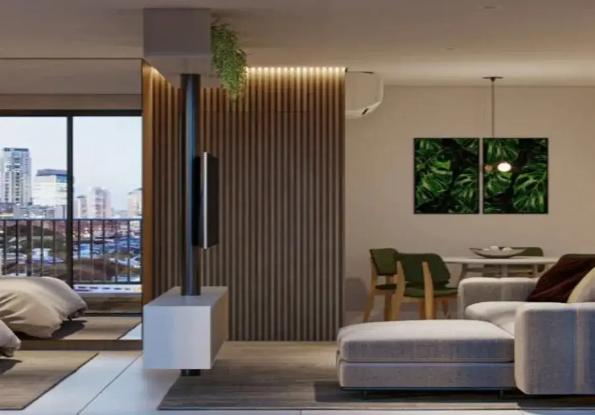 A imagem mostra o interior de um apartamento moderno, com um design de conceito aberto que integra sala de estar, sala de jantar e, parcialmente, o quarto. O ambiente geral é contemporâneo, funcional e aconchegante, com uma paleta de cores predominantemente neutra, pontuada por toques de cor e texturas naturais. Layout e Estrutura: O espaço é dividido de forma inteligente. Uma estrutura central funciona como divisor de ambientes e ponto focal: um pilar metálico escuro que vai do chão ao teto sustenta uma TV de tela plana. Abaixo da TV, um móvel baixo (rack/console) branco e flutuante está fixado ao pilar. Atrás dessa estrutura, um painel de madeira ripada na vertical, em tom de madeira médio, reveste parte da parede e se estende até próximo ao teto, onde uma iluminação embutida (provavelmente fita de LED) cria um efeito de luz indireta e acolhedora. No topo do painel, uma pequena planta pendente adiciona um toque de verde. Sala de Estar (Primeiro Plano): No primeiro plano, encontra-se a sala de estar. O móvel principal é um sofá modular (em L) estofado em tecido cinza claro texturizado, com almofadas que parecem ser em tons de vinho ou marrom escuro. O sofá repousa sobre um tapete retangular em tom de cinza escuro ou marrom, que delimita a área. O piso do apartamento parece ser de porcelanato ou vinílico em tom claro, possivelmente cinza bem suave ou bege. Sala de Jantar (Fundo à Direita): Ao fundo e à direita, parcialmente visível, está a área de jantar. Ela é composta por uma mesa de jantar branca e cadeiras com estrutura de madeira clara e assentos/encostos estofados em verde-oliva. Na parede acima da mesa, dois quadros formam um díptico com estampas de folhas tropicais grandes e verdes (como Costela-de-Adão), trazendo um elemento natural vibrante para o espaço. Uma luminária pendente simples, com uma lâmpada aparente, ilumina a mesa. A parede desta área é pintada em um tom neutro, como branco ou off-white. Um aparelho de ar condicionado split branco está instalado na parte superior da parede, acima do painel ripado. Quarto (Fundo à Esquerda): À esquerda, através de uma ampla abertura que leva a uma varanda, vislumbra-se parte do quarto. Uma cama com roupa de cama em tom neutro (bege ou taupe) está posicionada sobre um tapete escuro similar ao da sala. Varanda e Vista: A abertura para a varanda possui grandes portas de vidro, permitindo a entrada de luz natural e oferecendo uma vista urbana com prédios altos ao fundo. A varanda parece ter um guarda-corpo metálico. Cores e Materiais: A paleta de cores é baseada em neutros (cinza claro, cinza escuro, branco, bege, tons de madeira) com pontos de destaque em verde (plantas, quadros, cadeiras) e o metal escuro do pilar. Os materiais predominantes são madeira (painel ripado, cadeiras), metal (pilar, guarda-corpo), tecido (sofá, cadeiras) e vidro (portas da varanda). Ambiente: O ambiente transmite uma sensação de modernidade, organização e conforto urbano, com um design que valoriza a integração dos espaços, a funcionalidade e a incorporação de elementos naturais e boa iluminação.