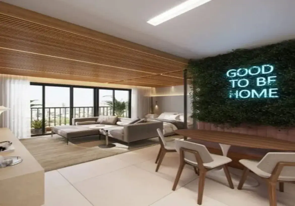 A imagem mostra o interior de um apartamento moderno, com um design de conceito aberto que integra sala de estar, sala de jantar e, ao fundo, uma área de dormir. Ambiente e Cores: O ambiente geral é sofisticado, acolhedor e contemporâneo. A paleta de cores principal é baseada em tons neutros e naturais: Neutros: O piso é revestido com grandes placas de porcelanato ou cerâmica em tom claro (off-white ou bege muito claro). As paredes são majoritariamente brancas ou em um tom neutro bem suave. O sofá é de um tom cinza-claro ou bege. Madeira: A madeira clara é um elemento de destaque, presente no teto ripado que cobre a área da sala e do quarto, e também na estrutura das cadeiras de jantar e no tampo da mesa. Isso traz calor e textura ao espaço. Verde: Um ponto focal vibrante é a parede da sala de jantar, revestida com um painel de folhagem verde (provavelmente artificial ou preservada), criando um efeito de jardim vertical. Azul Neon: Contrastando com o verde, um letreiro em neon azul claro com a frase "GOOD TO BE HOME" está instalado na parede de folhagem, adicionando um toque urbano e descolado. Rosa/Malva: Abaixo da parede verde, parece haver um banco estofado integrado à parede, num tom suave de rosa antigo ou malva, que serve de assento para a mesa de jantar. Móveis e Disposição: Sala de Estar: Dominada por um grande sofá secional em "L" com chaise, estofado em tecido cinza claro ou bege. Ele está posicionado sobre um tapete de cor neutra (bege/cinza) que delimita a área. Há uma pequena mesa de apoio redonda com tampo branco e base metálica. Ao fundo, grandes portas de vidro deslizantes com caixilharia preta dão acesso a uma varanda (com algumas plantas visíveis) e permitem a entrada de bastante luz natural. Cortinas brancas translúcidas estão recolhidas nas laterais. Em primeiro plano à esquerda, vê-se parte de um móvel baixo, provavelmente um rack ou aparador, em cor clara. Sala de Jantar: Localizada mais à direita, possui uma mesa de jantar com tampo de madeira de formato orgânico (possivelmente oval ou similar) e uma base central única e escultural na cor branca. Três cadeiras de design moderno são visíveis, com estrutura em madeira e assento/encosto estofados em tecido cinza claro ou bege. O banco estofado na parede complementa os assentos. Área de Dormir: Ao fundo, ligeiramente recuada e sob a continuação do teto de madeira ripada, está a área do quarto. É visível uma cama de casal com roupa de cama clara (branca e cinza/bege) e um criado-mudo com uma luminária. A iluminação parece ser indireta nesta área, vinda de uma sanca ou spots embutidos. Iluminação e Detalhes: Além da luz natural das portas da varanda, a iluminação artificial inclui uma luminária de teto retangular e alongada na parte branca do teto (sobre a área de jantar/circulação) e iluminação embutida ou indireta sobre a área da cama e possivelmente na transição entre o teto branco e o de madeira. O teto ripado de madeira é um detalhe arquitetônico marcante que unifica visualmente a sala e o quarto, conferindo aconchego. Em resumo, trata-se de um imóvel bem projetado, que utiliza cores neutras como base, aquecidas pela madeira, e adiciona pontos de interesse visual com a parede verde e o letreiro neon, criando um espaço funcional, moderno e convidativo.