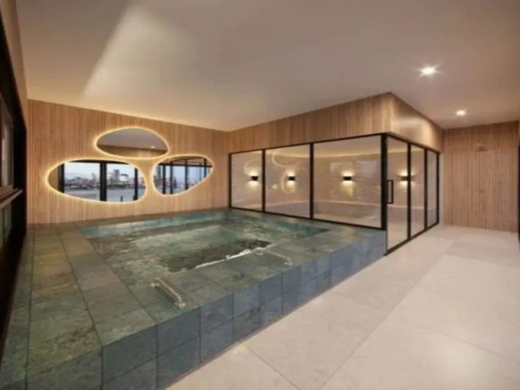 A imagem retrata um ambiente interno que parece ser uma área de spa ou piscina privativa, com um design moderno e sofisticado. Piscina: O elemento central é uma piscina retangular embutida no nível do chão. Ela é revestida com pastilhas ou azulejos quadrados de tamanho médio, em tons de verde-acinzentado escuro ou ardósia, que dão uma aparência profunda e natural à água. A água está calma, refletindo a iluminação do ambiente. Uma pequena escada metálica é visível dentro da piscina, próximo à borda esquerda. Paredes: As paredes predominantes são revestidas com painéis de madeira clara em formato de ripas verticais. Essa madeira confere uma sensação de calor e aconchego ao espaço, contrastando com a frieza da pedra da piscina e do vidro. Parede de Destaque (Esquerda): Uma das paredes laterais possui um design único com três grandes aberturas de formato orgânico e assimétrico (lembrando pedras ou amebas). Essas aberturas são contornadas por uma fita de LED embutida, que emite uma luz suave e quente, destacando seus contornos. Através dessas aberturas, é possível ver uma vista externa, aparentemente uma paisagem urbana com edifícios e água (rio ou mar), sugerindo que o imóvel está localizado em um andar alto com vista panorâmica. Parede de Vidro (Fundo/Direita): Adjacente à piscina, há uma grande divisória de vidro com esquadrias finas de metal preto. Essa estrutura cria um espaço separado, possivelmente uma sauna, sala de vapor ou área de relaxamento. Dentro deste espaço envidraçado, as paredes também são revestidas com a mesma madeira ripada, e há arandelas de parede emitindo luz quente para cima e para baixo. Piso: O piso ao redor da piscina é revestido com ladrilhos ou porcelanato de grande formato, em uma cor neutra, como bege claro ou off-white, com um acabamento que parece ser fosco ou levemente texturizado. Teto: O teto é liso e pintado de branco ou off-white. Possui iluminação embutida através de spots de luz estrategicamente posicionados, que fornecem a iluminação geral do ambiente. Há também um detalhe rebaixado ou inclinado no teto acima da área das janelas orgânicas. Cores e Materiais: A paleta de cores é predominantemente neutra e natural, combinando os tons quentes da madeira clara, o verde-acinzentado escuro da piscina, o bege claro do piso e o branco do teto. O preto das esquadrias de vidro e da janela lateral (parcialmente visível à esquerda) adiciona um contraste moderno. Os materiais principais são madeira, pedra/cerâmica (piscina e piso) e vidro. Ambiente e Iluminação: O ambiente geral é luxuoso, íntimo e relaxante. A iluminação desempenha um papel crucial, combinando a luz funcional dos spots no teto com a luz de destaque e ambiente das fitas de LED ao redor das aberturas e das arandelas dentro da área envidraçada. A iluminação é predominantemente quente, contribuindo para a atmosfera acolhedora. Móveis: Não há móveis tradicionais visíveis (como cadeiras, mesas ou espreguiçadeiras), reforçando a ideia de um espaço funcional focado na piscina e no relaxamento, possivelmente um spa residencial. Em resumo, a imagem mostra um espaço de piscina interna elegante e contemporâneo, caracterizado pelo uso de materiais naturais como madeira e pedra, um design de iluminação cuidadosamente planejado, uma paleta de cores sóbria e uma vista externa através de janelas com design orgânico e iluminado.