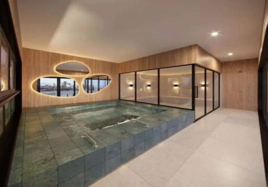 A imagem retrata um ambiente interno que parece ser uma área de spa ou piscina privativa, com um design moderno e sofisticado. Piscina: O elemento central é uma piscina retangular embutida no nível do chão. Ela é revestida com pastilhas ou azulejos quadrados de tamanho médio, em tons de verde-acinzentado escuro ou ardósia, que dão uma aparência profunda e natural à água. A água está calma, refletindo a iluminação do ambiente. Uma pequena escada metálica é visível dentro da piscina, próximo à borda esquerda. Paredes: As paredes predominantes são revestidas com painéis de madeira clara em formato de ripas verticais. Essa madeira confere uma sensação de calor e aconchego ao espaço, contrastando com a frieza da pedra da piscina e do vidro. Parede de Destaque (Esquerda): Uma das paredes laterais possui um design único com três grandes aberturas de formato orgânico e assimétrico (lembrando pedras ou amebas). Essas aberturas são contornadas por uma fita de LED embutida, que emite uma luz suave e quente, destacando seus contornos. Através dessas aberturas, é possível ver uma vista externa, aparentemente uma paisagem urbana com edifícios e água (rio ou mar), sugerindo que o imóvel está localizado em um andar alto com vista panorâmica. Parede de Vidro (Fundo/Direita): Adjacente à piscina, há uma grande divisória de vidro com esquadrias finas de metal preto. Essa estrutura cria um espaço separado, possivelmente uma sauna, sala de vapor ou área de relaxamento. Dentro deste espaço envidraçado, as paredes também são revestidas com a mesma madeira ripada, e há arandelas de parede emitindo luz quente para cima e para baixo. Piso: O piso ao redor da piscina é revestido com ladrilhos ou porcelanato de grande formato, em uma cor neutra, como bege claro ou off-white, com um acabamento que parece ser fosco ou levemente texturizado. Teto: O teto é liso e pintado de branco ou off-white. Possui iluminação embutida através de spots de luz estrategicamente posicionados, que fornecem a iluminação geral do ambiente. Há também um detalhe rebaixado ou inclinado no teto acima da área das janelas orgânicas. Cores e Materiais: A paleta de cores é predominantemente neutra e natural, combinando os tons quentes da madeira clara, o verde-acinzentado escuro da piscina, o bege claro do piso e o branco do teto. O preto das esquadrias de vidro e da janela lateral (parcialmente visível à esquerda) adiciona um contraste moderno. Os materiais principais são madeira, pedra/cerâmica (piscina e piso) e vidro. Ambiente e Iluminação: O ambiente geral é luxuoso, íntimo e relaxante. A iluminação desempenha um papel crucial, combinando a luz funcional dos spots no teto com a luz de destaque e ambiente das fitas de LED ao redor das aberturas e das arandelas dentro da área envidraçada. A iluminação é predominantemente quente, contribuindo para a atmosfera acolhedora. Móveis: Não há móveis tradicionais visíveis (como cadeiras, mesas ou espreguiçadeiras), reforçando a ideia de um espaço funcional focado na piscina e no relaxamento, possivelmente um spa residencial. Em resumo, a imagem mostra um espaço de piscina interna elegante e contemporâneo, caracterizado pelo uso de materiais naturais como madeira e pedra, um design de iluminação cuidadosamente planejado, uma paleta de cores sóbria e uma vista externa através de janelas com design orgânico e iluminado.