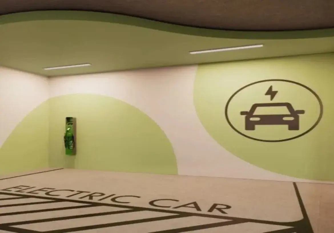 A imagem mostra uma vaga de estacionamento interna, especificamente designada para o carregamento de veículos elétricos. O espaço é bem iluminado por duas luminárias de teto longas e retangulares, de luz branca. O piso da vaga é de cor clara, possivelmente concreto polido ou epóxi cinza claro, e possui marcações proeminentes em cinza escuro ou preto. Na parte mais próxima do observador, lê-se em letras maiúsculas "ELECTRIC CAR". Abaixo do texto, faixas diagonais preenchem a área da vaga, delimitando claramente o espaço reservado. As paredes são predominantemente brancas, mas decoradas com grandes formas curvas pintadas em um tom de verde claro, quase pistache. Na parede do fundo, à esquerda, está instalada a Estação de Recarga para Veículos Elétricos. É um equipamento vertical, de cor verde vibrante, montado na parede, com o que parece ser um cabo de carregamento enrolado ou pendurado na sua frente. Na parede da direita, uma grande forma circular em verde claro serve de fundo para um símbolo universal de carregamento de veículos elétricos: um ícone estilizado de um carro (visto de frente) com um símbolo de raio (eletricidade) acima dele, tudo dentro de um círculo, em cor cinza escuro ou preto. O teto é predominantemente branco, mas acompanha a parede do fundo com uma faixa curva rebaixada ou saliente, também pintada em verde claro, que segue a linha ondulada da decoração da parede, conferindo um design moderno e coeso ao ambiente. A atmosfera geral é limpa, funcional e claramente sinalizada para sua finalidade. Palavra chave: Apartamento com 2 Quartos a Venda Azure Compact Life Marista
