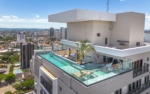 Vista aérea da área da piscina no topo do prédio, mostrando o deck, palmeira decorativa e a densidade urbana ao fundo.