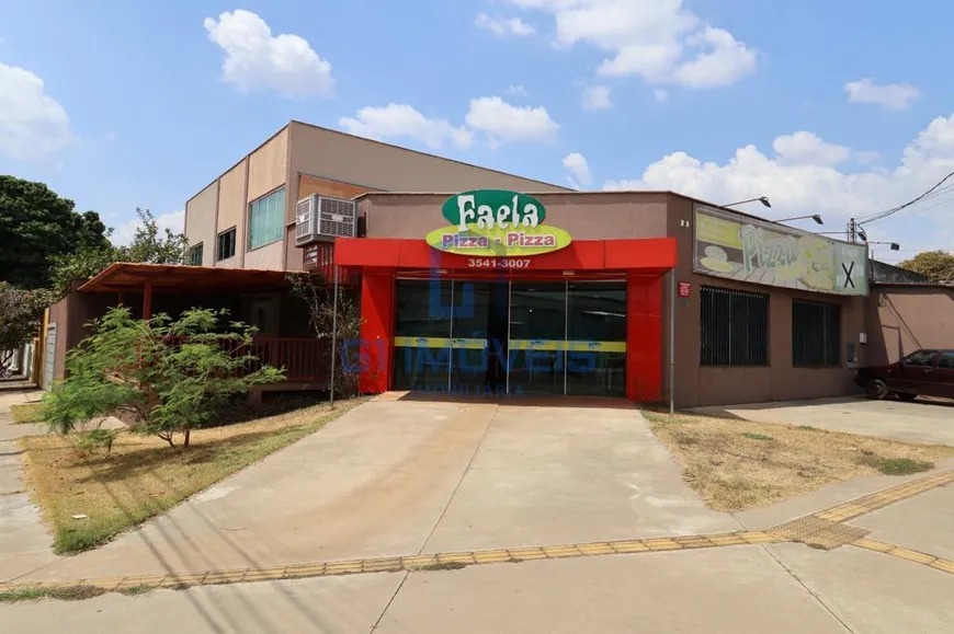 Vista lateral da pizzaria mostrando a entrada principal vermelha, calçada ampla e estacionamento.